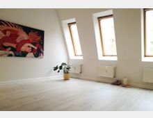 Ein minimalistischer Innenraum mit hellem Holzboden, weißen Wänden und drei großen Dachfenstern, die natürliches Licht hereinlassen. An der Wand hängt ein abstraktes Kunstwerk, daneben steht eine grüne Zimmerpflanze in einem Korb, und unter einem Fenster liegen eine Kerze und ein Tablett auf einem kleinen Teppich.