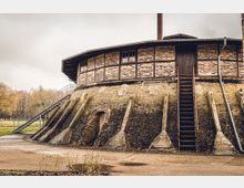 Rundbau aus Backstein mit Fachwerk-Elementen, seitlich Treppenaufgänge und am unteren Bereich gestützte Bögen, umgeben von einer Parklandschaft mit kahlen Bäumen. Ein hoher Schornstein ragt aus der Mitte des Gebäudes hervor.
