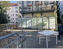 Ein offener Hofbereich mit gepflastertem Boden, einem weißen Tisch aus Kunststoff und Plastikstühlen. Im Hintergrund sind Balkone eines Mehrfamilienhauses und eine halbtransparente Sichtschutzwand aus Glas zu sehen, umgeben von Pflanzen und Bäumen.