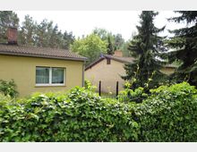 Zwei eingeschossige Wohnhäuser mit Satteldächern, umgeben von Bäumen und einer dichten Hecke aus efeubewachsenen Pflanzen im Vordergrund. Im Hintergrund sind hohe Nadelbäume und ein Zaun zu sehen.