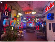 Ein Innenraum mit zahlreichen bunten, leuchtenden Buchstaben und Schriftzügen an den Wänden, Regalen und Decken. Der Raum hat eine industrielle Ästhetik mit unverputzten Wänden, Metallregalen und gedecktem Licht.