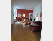 Wohnbereich mit Parkettboden im Fischgrätmuster, einer roten Wand, einem großen Esstisch mit gemischten Stühlen und einem goldgerahmten Spiegel. Im Vordergrund steht ein rotes Ecksofa neben einem niedrigen Couchtisch auf einem Teppich.