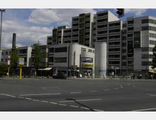 Mehrstöckiges modernes Wohn- und Geschäftshaus mit grauer Fassade und vereinzelt gelben Akzenten, vor einem großen Straßenkreuzungspunkt. Im Vordergrund sind Schilder für die U-Bahn-Station „U-Bhf Richard-Wagner-Platz“ und Geschäfte im Erdgeschoss sichtbar.