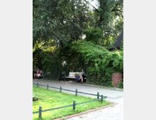 Ein kleiner Park mit einer gepflasterten Gehwegfläche und grüner Rasenfläche im Vordergrund. Im Hintergrund sind Holzbänke unter einer rankenden Laubenpergola aus dichtem Efeu zu sehen, die von hohen Bäumen umgeben ist. Links sitzen Personen auf den Bänken.