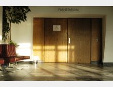 Eingangsbereich eines Planetariums mit einer Holz-Doppeltür in der Mitte, beschriftet mit "Planetarium" über dem Türrahmen. Links im Bild stehen eine rote Polsterbank und eine grüne Zimmerpflanze, der Boden besteht aus hellen Steinfliesen.
