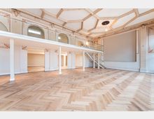 Heller, leerer Saal mit großem Fischgrät-Parkett und einer umlaufenden Empore mit Glasgeländer und Treppe. Die hohe, reich verzierte Kassettendecke sowie Rundbögen und Wandstuck prägen den historischen Innenraum.