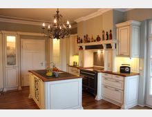 Klassische Küche mit cremefarbenen Einbauschränken, einer zentralen Kochinsel mit Holzarbeitsplatte und integriertem Weinschrank. Ein großer schwarzer Herd mit Dunstabzugshaube, ein dekorativer Kronleuchter und ein Holzboden prägen den Raum, der durch Deckenelemente und eingelassene Beleuchtung ergänzt wird.