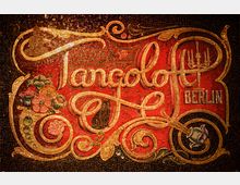 Mosaik-Wandbild mit goldenen und roten Elementen zeigt den Schriftzug „Tangoloft Berlin“ in verzierter, geschwungener Schrift. Ergänzt wird das Design durch florale Muster, Ornamente und einen Kronleuchter.