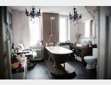 Ein Badezimmer mit einer freistehenden Badewanne auf einem Podest aus Mosaikfliesen, flankiert von zwei Fenstern. Der Raum enthält schwarze Kronleuchter, einen Waschtisch mit Spiegel sowie eine Toilette, umgeben von zahlreichen Pflegeprodukten und Dekorationen.