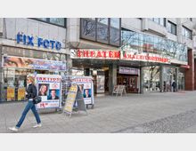 Straßenansicht eines Gebäudes mit gläserner Fassade, einem Fotogeschäft namens "Fix Foto" links und dem Eingang zum "Theater am Kurfürstendamm" rechts, gekennzeichnet durch ein rotes Leuchtschild. Im Vordergrund stehen Werbeschilder für Bewerbungsfotos und eine Frau geht auf dem Gehweg vorbei.