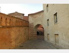 Gasse mit Kopfsteinpflaster, flankiert von einer hohen roten Backsteinmauer auf der linken Seite und einer hell verputzten Hausfassade mit kleinen Fenstern auf der rechten Seite; im Hintergrund ein gemauerter Torbogen, der zu einem Innenhof führt.