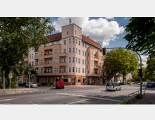 Ecke eines Wohn- und Geschäftshauses mit heller Fassade und orangen Akzenten, vier Stockwerken und einem Mansardendach, an einer Kreuzung mit Ampeln, parkenden Autos und umgebenden Bäumen.