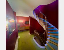 Treppenhaus in einem Innenraum mit geschwungener Treppe, goldfarbenem Geländer und grünem Teppichboden. Die Wände sind rot gestrichen, dekoriert mit einem Wandspiegel, einer Kommode und Wandleuchten.