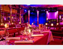 Ein festlich gedeckter Veranstaltungsraum mit rot eingedeckten Tischen, Kerzenlicht und zahlreichen Gläsern; im Hintergrund eine Bühne mit blau beleuchteten Vorhängen, Holzverzierungen und einem Klavier.