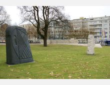 Grünfläche mit zwei modernen Skulpturen, eine aus dunklem Stein mit geometrischen Mustern und eine aus hellem Stein mit Reliefmotiven, im Hintergrund Bäume und mehrstöckige Wohn- und Geschäftshäuser.