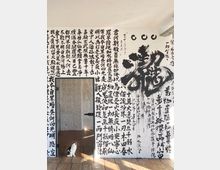 Weiße Wand mit schwarzer, handgemalter Kalligrafie in chinesischen Schriftzeichen; zentrale große Schrift trägt eine expressive Pinselästhetik. Eine Tür in der Wand öffnet sich in einen angrenzenden Raum mit weißem Mauerwerk; auf dem Holzboden sitzt eine schwarz-weiße Katze.