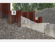 Ein Lagerplatz mit Kieshaufen, getrennt durch rostfarbene Metalltrennwände, die mit den Nummern 9 und 10 gekennzeichnet sind. Im Hintergrund sind Bäume und weitere Schotterhaufen sichtbar.