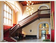 Historisches Treppenhaus mit einer hölzernen, verzierten Treppe, rotem Teppich und einem hohen Fenster, das viel Licht einlässt. Im Erdgeschoss befinden sich schwarze Sitzmöbel, ein Gemälde an der Wand sowie dekorative Elemente, und eine prächtige Deckenleuchte hängt im Zentrum des Raums.
