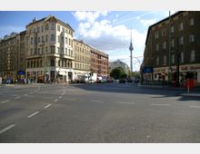 Straßenszene an einer belebten Kreuzung in Berlin mit mehrstöckigen Altbauten, Geschäften im Erdgeschoss und einem U-Bahn-Eingang mit der Beschriftung „Alexanderplatz“. Im Hintergrund ragt der Berliner Fernsehturm in den Himmel.