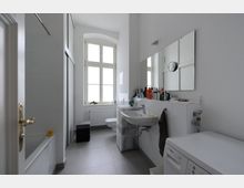 Helles Badezimmer mit weiß gefliesten Wänden, einer Badewanne links, einem Waschbecken und einem großen Spiegel rechts. Im Hintergrund befindet sich ein großes Fenster mit eingelassenem Heizkörper, daneben eine Waschmaschine und ein Hocker.