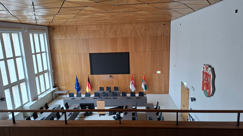 Moderner Ratssaal mit U-förmig angeordneten Tischen und Mikrofonplätzen vor einer holzverkleideten Stirnwand mit großem Bildschirm und Fahnen. Links sorgen hohe Fenster für Tageslicht, darüber spannt sich eine geometrische Holzdecke; an der rechten Wand hängt ein Wappen.