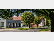 Moderne Rettungswache mit einer schlichten grauen Fassade und großen Toren, die von Bäumen und einer ruhigen Straße mit Gehweg umgeben ist. Im Hintergrund sind Wohngebäude mit roten Ziegeldächern zu sehen.