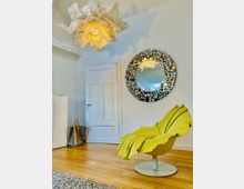 Helles Wohnzimmer mit einer weißen Tür, einem großen, runden Spiegel aus metallischen Akzenten und einer gelben Designer-Stuhl in Blattform. Eine auffällige, skulpturale Deckenleuchte mit einem Stoffdesign hängt an der weißen Decke, während der Holzboden und ein beiger Teppich den Raum ergänzen.