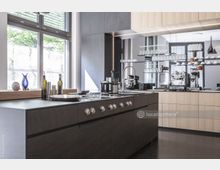 Moderne Küche mit einer großen Kochinsel aus dunklem Holz und Edelstahl-Elementen, flankiert von hellen Holzschränken. Im Hintergrund befinden sich große Fenster, Regale mit Gläsern und Küchenutensilien sowie eine Kaffeemaschine.