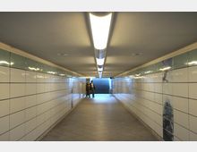 Ein beleuchteter unterirdischer Fußgängertunnel mit weißen, glänzenden Wandfliesen und einer langen, zentralen Deckenleuchte. Am Ende des Tunnels führen Treppenstufen nach oben, daneben stehen zwei Personen.
