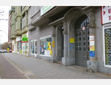 Straßenansicht mit einer Häuserzeile, bestehend aus einem grauen Gebäude mit verzierten Rundbögen und angrenzenden Geschäften. Vor den Schaufenstern befinden sich Werbeplakate, während auf dem Gehweg einige Schilder und ein Aufsteller zu sehen sind.