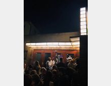 Außenansicht eines Kinos bei Nacht mit leuchtendem Schriftzug "KINO" über dem Eingang. Vor der Tür aus dunkelbraunem Holz steht eine Menschenmenge, die sich unterhält, umgeben von gedämpftem Licht.