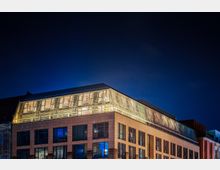 Moderne Bürogebäude bei Nacht mit rechteckigen Fassadenflächen aus Glas und Stein sowie einem beleuchteten Obergeschoss mit Lamellenstruktur. Der Himmel im Hintergrund ist dunkelblau und klar.