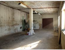 Ein leerer, einfacher Raum mit rohem Betonboden, unverputzten Wänden und einer freiliegenden Betonsäule in der Mitte. Im Hintergrund befinden sich ein großes Holztor und einige verstreute Gegenstände; Tageslicht scheint durch ein Fenster auf der rechten Seite herein.