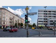 Straßenkreuzung mit Mehrfamilienhäusern auf beiden Seiten; links ältere Gebäude mit verzierten Fassaden, rechts ein modernes Wohn- und Geschäftshaus. Im Vordergrund eine Ampel mit grünem Signal, Verkehr inklusive Autos und einem gelben Bus, sowie Straßenschilder, darunter eines mit der Aufschrift „Kaiser-Friedrich-Straße“.