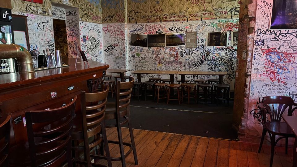 Innenraum einer Kneipe mit dunkler Holztheke und Barhockern auf Holzdielenboden. Die Wände und die Decke sind dicht mit Graffiti und Kritzeleien bedeckt, dazu einfache Tische und Stühle sowie eine kleine erhöhte Sitzecke an der Rückwand.