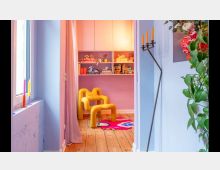 Ein farbenfrohes Zimmer mit pastellblauen und rosa Wänden, einem gelben Designerstuhl, einem Teppich mit kräftigen Mustern und Holzfußboden. Im Hintergrund befinden sich Regale mit Spielzeug und Büchern, während im Vordergrund Pflanzen, Kerzen und ein schwarzer Kerzenständer zu sehen sind.