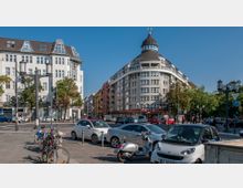 Straßenszene mit einem markanten, halbrunden Gebäude mit Kuppeldach und modernen Fenstern im Hintergrund. Im Vordergrund sind parkende Autos, Fahrräder und Motorroller sowie mehrere Straßenlaternen und Ampeln zu sehen; um das Gebäude herum stehen Bäume.