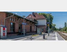 Bahnhofsgebäude des Bahnhofs "Beelitz Stadt" mit rotem Ziegelmauerwerk und Satteldach. Daneben ein moderner Bahnsteig mit Überdachung, Sitzmöglichkeiten, einem Fahrkartenautomaten und Gleisen im Grünen.