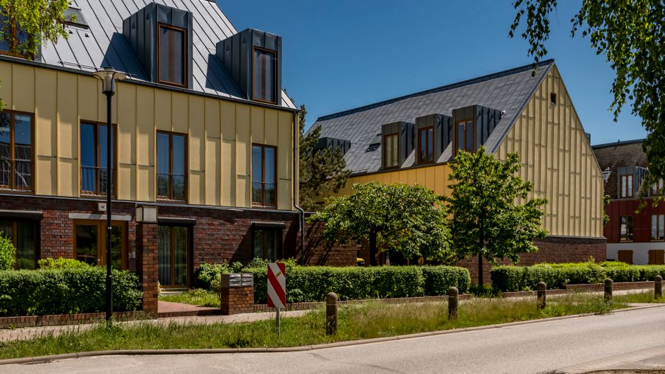 Moderne Gebäudezeile an einer Straße mit hellen Fassaden über einem rotbraunen Ziegelsockel und geneigten Metalldächern mit Gaubenfenstern. Davor liegen Hecken und kleine Bäume entlang des Gehwegs, eingerahmt von Straßenlaternen und einem Baum am rechten Bildrand.
