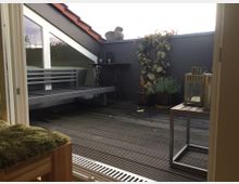 Kleine Dachterrasse mit Holzboden, flankiert von einer dunklen Brüstung und einem schrägen Fenster. Auf der Terrasse stehen ein graues Sitzmöbel, Pflanzen in Töpfen und eine kleine Metalllaterne auf einem Beistelltisch.