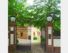 Eingangstor zu einem Grundstück mit Backsteinpfeilern und schwarzen Metallgittern, verziert mit weißen Lampen. Im Hintergrund ist ein großzügiges, gelbes Gebäude mit einem Balkon und umgeben von Bäumen und gepflegtem Garten zu sehen.