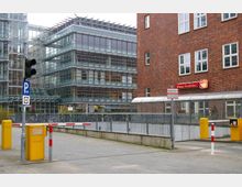 Eine städtische Szene mit einem modernen Bürogebäude aus Glas und Stahl links und einer Fassade aus rotem Ziegelstein rechts. Im Vordergrund befinden sich eine Schranke, gelbe Parkautomaten und ein Verkehrsschild für Parkplätze.