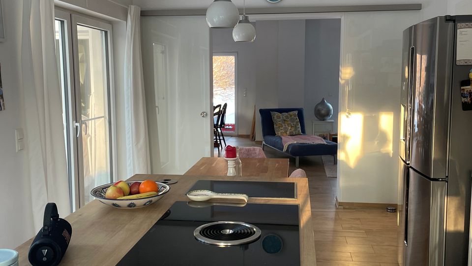 Moderne, helle Wohnküche mit großer Holz-Kücheninsel und integriertem schwarzem Kochfeld, darüber hängende Kugel-Pendelleuchten und eingelassene Deckenstrahler. Links führen große Glas-Schiebetüren mit Vorhängen nach außen, rechts stehen hohe weiße Fronten und ein Edelstahl-Kühlschrank; im Hintergrund öffnet sich der Raum zu einem Wohnbereich.