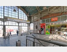 Eingangsbereich eines Bahnhofs mit großen Glasfenstern, sichtbaren Metallträgern und einem grünen Wellblechdach. Rechts befindet sich ein kleiner Laden mit Snacks und Getränken sowie ein Geldautomat; links führt ein Ausgang zur Straße.