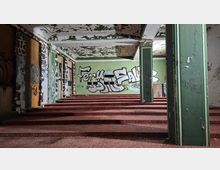 Innenraum eines verlassenen Gebäudes mit breiten, rotbraunen Stufen, grünen Wänden und mehreren Stützpfeilern. Abblätternde Deckenfarbe, teils zugemauerte Fenster und großflächige Graffiti prägen den Raum.
