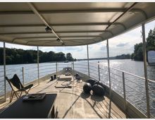 Sicht von einem überdachten Deck eines Bootes mit Geländern an beiden Seiten, Blick auf eine ruhige Flusslandschaft mit Bäumen am Ufer und Sonnenreflexionen auf dem Wasser. Auf dem Deck befinden sich ein Stuhl, eine Tischplatte und mehrere Fender.