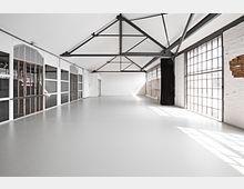 Offener Industrie-Loftraum mit weiß gestrichenen Wänden, großen Gitterfenstern, freiliegenden Stahlträgern in der Decke und einem glatten, hellen Boden. Auf der linken Seite befinden sich Fenster mit Holzrahmen, rechts gemauerte Wände mit partiell freigelegtem Ziegelwerk.