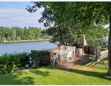 Terrasse aus Holz mit Blick auf einen See, umgeben von Bäumen und Sträuchern. Auf der Terrasse stehen weiße Gartenmöbel und eine Schaukelbank; im Hintergrund ist eine bewaldete Uferlandschaft sichtbar.