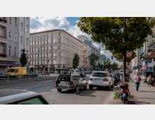 Städtische Straßenszene mit mehrstöckigen, historischen und modernen Gebäuden, parkenden Autos und einem Baum am Straßenrand. Fußgänger, geöffnete Geschäfte und Cafés beleben den Gehweg, während Fahrzeuge die mehrspurige Straße befahren.