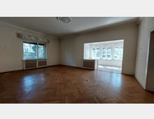 Großer, leerer Raum mit Parkettboden und weiß gestrichenen Wänden, einem großen Fenster auf der linken Seite und einem angrenzenden Wintergarten mit zahlreichen Fenstern, die Tageslicht hereinlassen. Die Decke ist mit einer dekorativen Rosette versehen, und unter den Fenstern befinden sich Heizkörper.